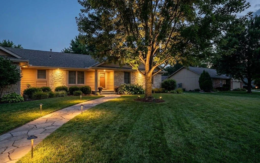 Landscape lighting installed along the front walkway and garden beds of a newer single-family home in Maize, Kansas, with warm light accenting the home's elevation after dark. Low-profile fixtures illuminate a tree line and driveway edge with no visible glare.